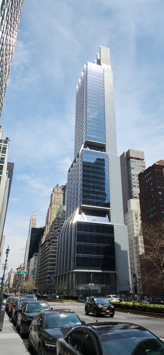 Tall shiny building, silver and white. Tapers and has three parts, could be a spaceship! Blue sky light clouds, large street with cars in foreground, Park Avenue. NYC