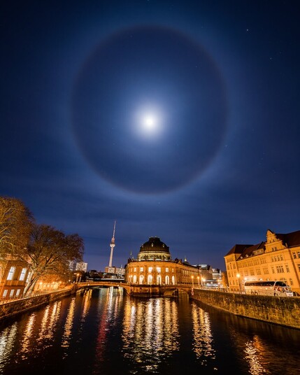 The moon at the centre, with a large, misty circle in the sky around it, above some famous Berlin landmarks, the TV tower and Museum Island.
