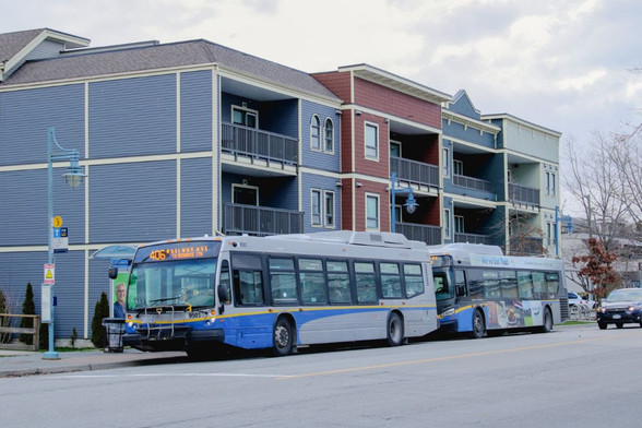 Two single deck buses are waiting  at a bus stop. Behind them is a three storey building but the ground floor cannot be seen due to the buses presence

Translink photo