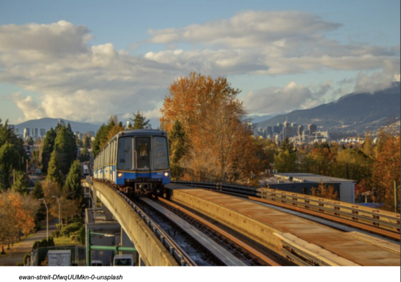 Translink Mark I SkyTrain in Black and Blue livery approaching the camera on an elevated track. The background is an urban area with trees turning brown and distant high-rises and the North Shore mountains.  