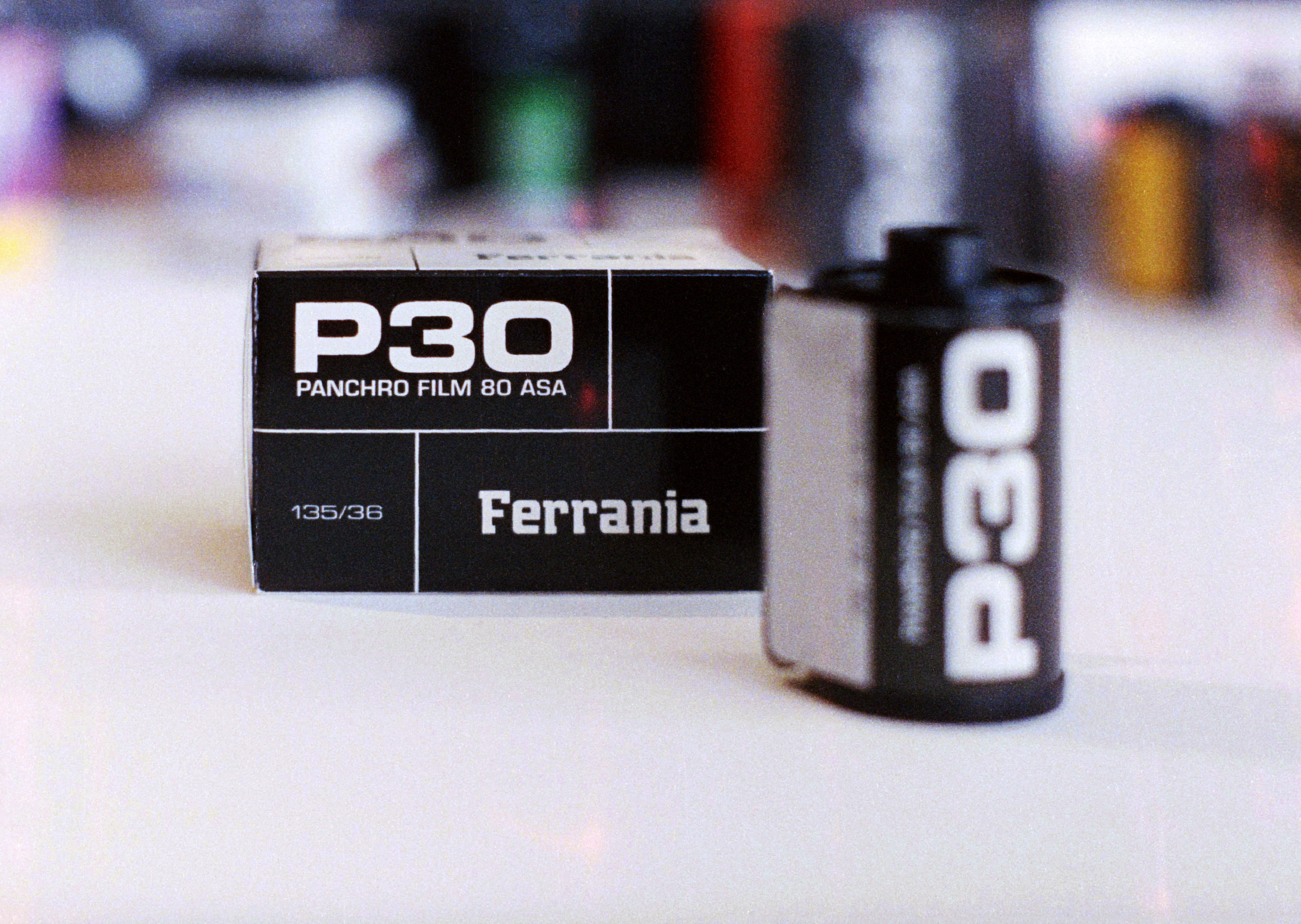 A box of Film Ferrania P30 film next to 35mm cartridge on a white table with some colourful out-of-focus background.
