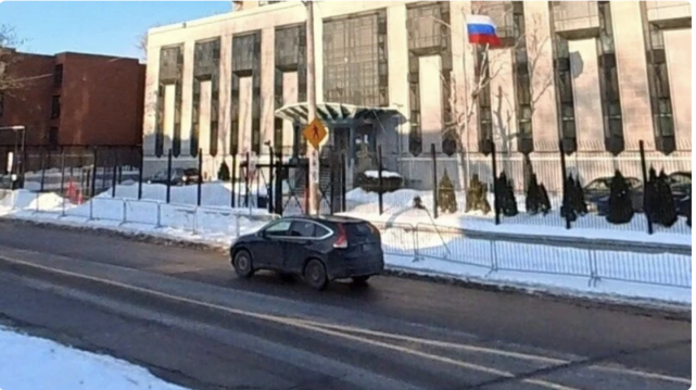 A grim flat fronted concrete building with a Russian flag flying outside. There is a lot of snow about but the street has been cleared. Just one SUV is using the street in the picture. No other vehicle of person is visible