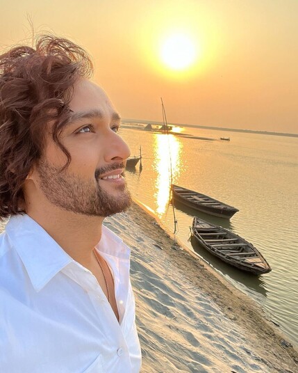 Sourabh Raaj Jain stands on the sandy banks of the Saryu river, near Gola Bazar in Uttar Pradesh, with the bright yellow early morning sun low in the amber sky behind him and its golden light reflecting on the surface of the gently rippling water. Fishing boats are moored nearby, and the distant opposite bank of the river can be seen on the horizon. Sourabh is to the left of the frame, looking up at the sky as though looking at the sun with it in front of him. His mid-length, warm brown hair is curled for the shoot and his beard and moustache are a little longer than usual. He wears a crisp, plain white shirt, a silver chain visible within the collar.