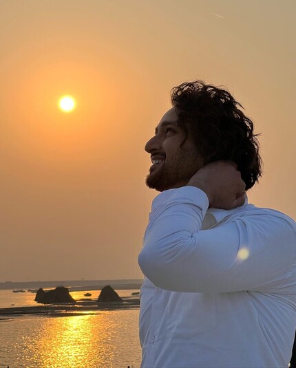 Sourabh Raaj Jain stands beside the Saryu river, the evening sun low in the sky behind him and its golden light reflecting on the surface of the gently rippling water. The distant opposite bank of the river can be seen on the horizon. Sourabh is to the right of the frame, looking up at the sky as though looking at the sun and with his left hand resting on his neck. 