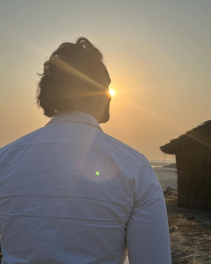 Sourabh Raaj Jain is seen from behind walking towards the Saryu river, the edge of his right cheek partially obscuring the evening sun low in the sky in front of him. He is on the left of the frame. In front of him and to the right is a small grass hut.  