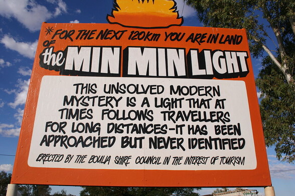 This is a photo of a street sign in Australia.

It says:

"For the next 120km you are in land of the Min Min Light.

This unsolved modern mystery is a light that at times follows travellers for long distances - it has been approached but never identified.

Erected by the Boula Shore Council in the interest of tourism."

The sign is bright orange and the background shows trees and the sky.