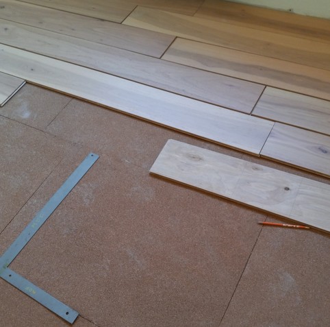 Hardwood flooring planks loosely laid out over top of cork underflooring along with a yellow pencil and a dull gray square ruler.