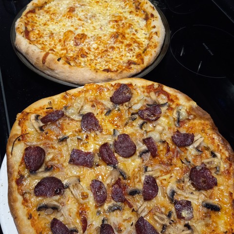Two homemade pizzas - cheese and dry sausage, onions, mushrooms, and cheese - on a black stove top. 
