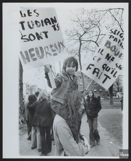 Black-and-white photograph showing university students marching in the background and, in the foreground, a long-haired student carrying a young boy on his shoulders. The boy is holding two handwritten protest signs, on one is written "Les étudiants sont heureux" [the students are happy] and on the other "Seigneur pardon Bour[? Bourassa?] il ne sait ce qu'il fait" [Lord forgive Bour[? Bourassa?] for he knows not what he does].