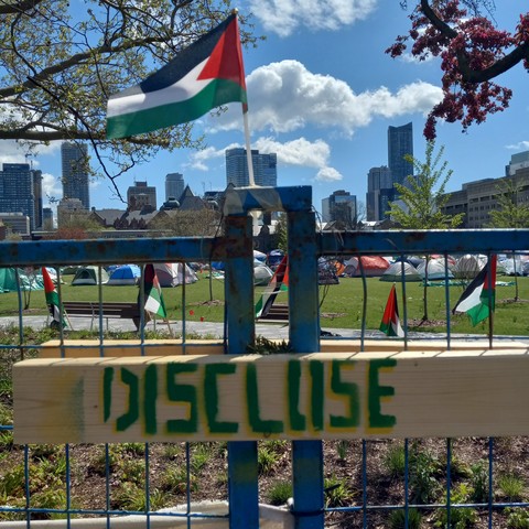 "DISCLOSE" stencilled in green and yellow on a two by four holding two sections of construction fencing together. Above floats a Palestinian flag. Through the fence, we see the student encampment and the Toronto skyline in the distance.