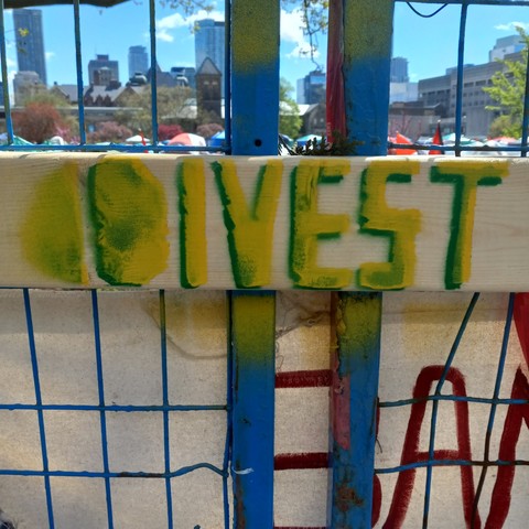 "DIVEST" stencilled in yellow and green on a two by four holding two sections of construction fencing together. Through the fence, we see the student encampment and the Toronto skyline in the distance.