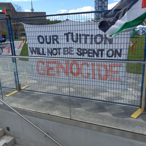 A white banner with black and red lettering hanging on blue construction fencing: "OUR TUITION WILL NOT BE SPENT ON GENOCIDE". The banner is flanked by two Palestinian flags.