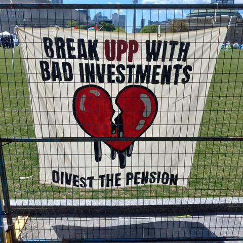 A white banner with black and red lettering featuring a red broken heart with black ink dripping from it hanging on blue construction fencing: "BREAK UPP WITH BAD INVESTMENTS. DIVEST THE PENSION". The UPP is the University Pension Plan, made up of the University of Toronto and other Ontario universities.
