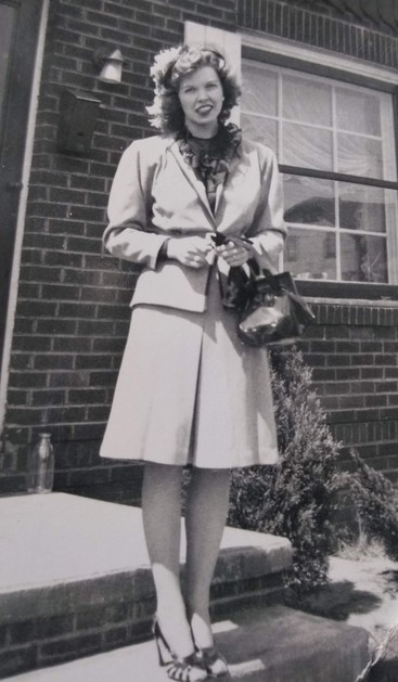 In mid 1940s a well dressed young woman stands on front steps of a brick house.  She is wearing typical clothing of the time, a light colored suit with shoulder pads and slightly flared skirt cut just above the knee. Her blouse is darker, ruffled at the neck. She wears open toed high heeled sandals. Holds a handbag in front of her as she smiles for the camera.