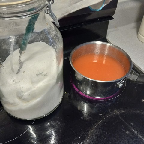 Reddish liquid in a small kettle next to a large jar full of refined sugar. 