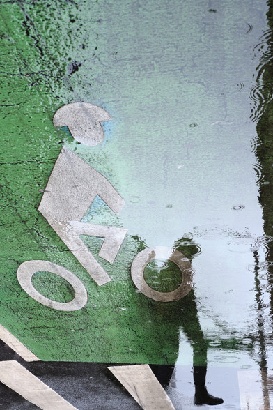 A puddle blending a bike lane painted sign with a distorted silhouette  in the water about to take a step.

The person is distorted and you can only make out their silhouette. The surface of the water is uneven and covered with ripples from raindrops. The person's foot is the least distorted, it hangs out in front of the person as they are about to take a step.

At the bottom of the image there are large crosswalk stripes.