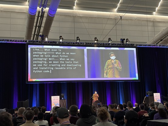 Sumana on stage with blue and purple lighting. The transcribed talk behind her says "Like... What even is packaging??????" with six question marks.