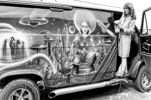 A white woman with long curly blond hair leaning out of the front passenger door of a boogie van, inspecting its custom science-fiction-themed, airbrushed paint job.