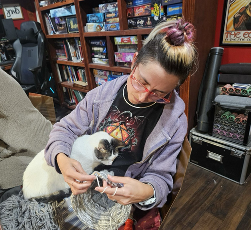 A woman in glasses leaned over looking at her knitting while a white and tabby pointed cat is curled under her arm, also looking at the knitting 