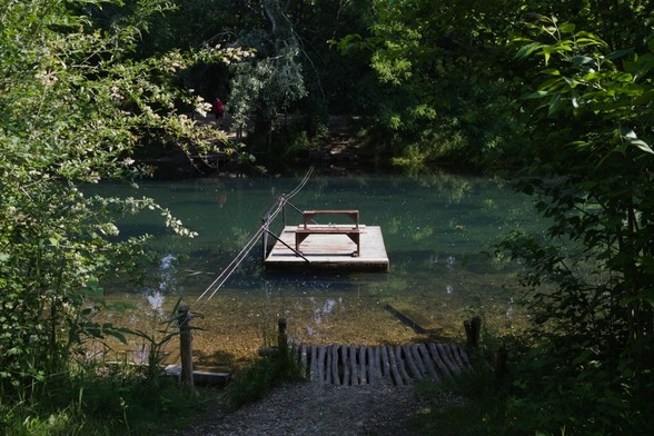Eine kleine handbetriebene Fähre mit 2 Sitzbänken. Die Fähre hängt am Seil mitten im Fluss. Es führt ein kleiner Weg durch die Bäume zur Anlegestelle.