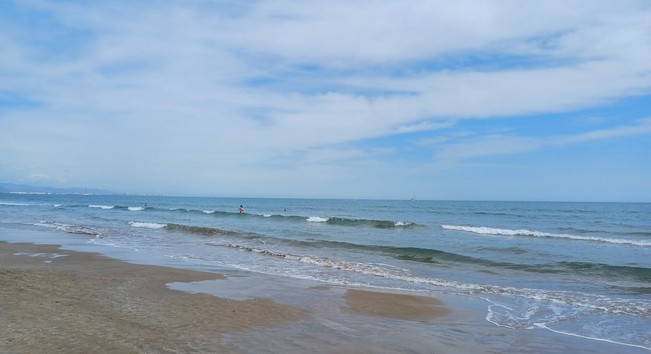 A serene beach scene with the ocean meeting the sky at the horizon. The sky is partly cloudy, suggesting a pleasant day. The ocean has gentle waves, and there are a few people in the water, enjoying the surf or swimming. The beach us sandy, and the tide is coming in, as indicated by the water reaching further up the shore. The overall atmosphere is calm and inviting, typical of a beach setting.