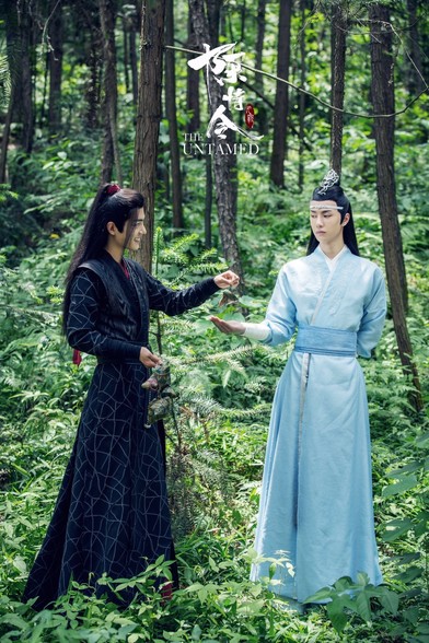 Lan Wangji (in light blue robes) holding out his hand for Wei Wuxian (in black robes) who is dangling a zongzi above his hand. He has several more zongzi in his other hand. 