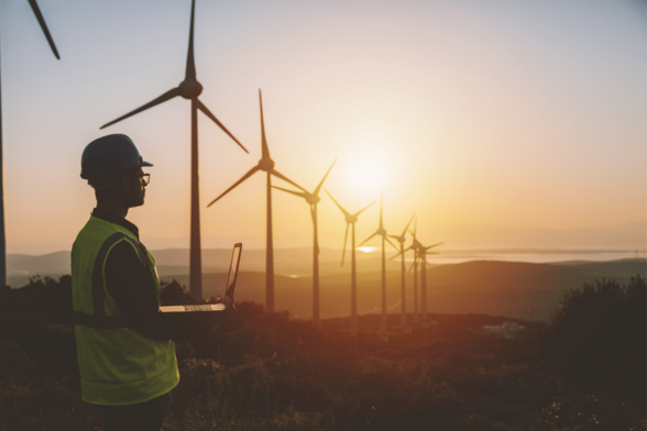 Nine wind turbines - a shot taken into the sun - with a man wearing a hi-vis vest and a helmet while holding an open laptop in his hands