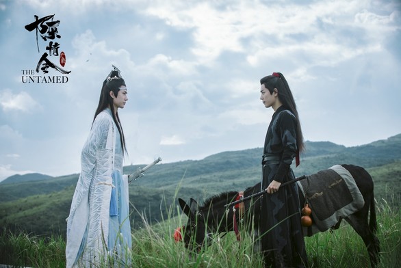 Lan Wangji (in white and light blue robes) and Wei Wuxian (in black robes) with Little Apple standing on a green hill opposite to each other