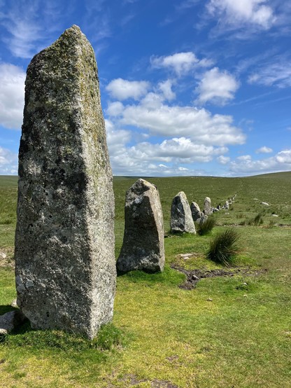 Photo taken just in front of the leading stone pillar. It’s angular, the straight sides crusted with yellow and grey lichens. Stones lead away from it to the right, in decreasing height initially. 
A few tufts of dark green soft rush sprout up from the short turf either side of the row.
Wispy white clouds scud across the blue sky.