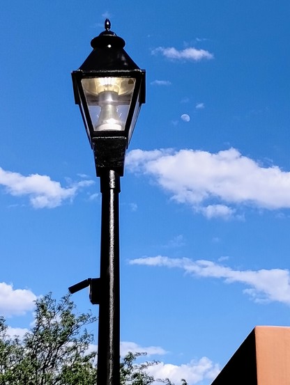 The top of a tall, black lamppost with an enclosed rectangular light sits in front of a blue sky with some scattered clouds. A small, crescent moon sits to the right of the light. It floats above a few clouds and almost looks like a cloud itself. The top of a full, green tree peaks out from the lower left corner behind the lamppost. The top corner of a building fills the lower, right corner.