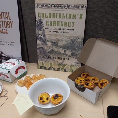 The blown-up cover of a book (Colonialism's Currency: Money, State, and First Nations in Canada, 1820-1950) with a bowl and a pastry box, both holding pastel de nata (Portuguese custard tarts).