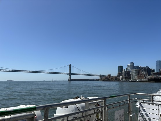 Clear bright blue sky and blue water. The Golden Gate Bridge entering San Francisco with its tall buildings. The view is from the back of a ferry on the water, with some of its deck and railing in the foreground. 