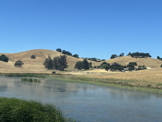 Blue sky, golden brown hills with green trees. A blue river with green banks in the foreground. A hill on the right begins to obscure some buildings including a large barn. 