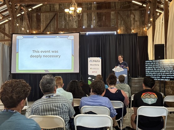 Chris holding a mic behind a lectern on a stage in a barn. The screen shows a slide that says “This event was deeply necessary.”