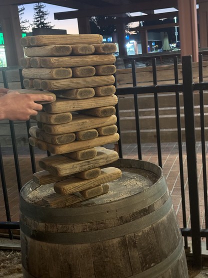 Large wooden blocks precariously stacked on top of each other. Hands are removing one from the middle of the stack. 