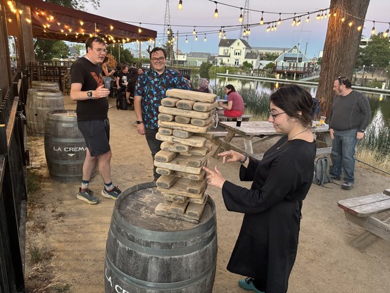 A person in a black dress carefully tests removing a wooden block from a Jenga stack. Others look on in trepidation.