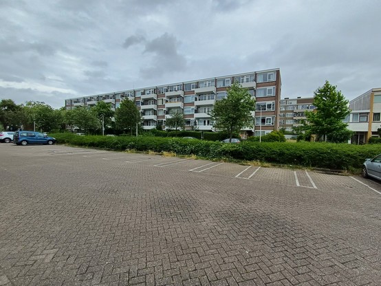 Wide angle picture of a parking space that is mostly empty, with a 5 story flat in the background, under a grey cloudy sky