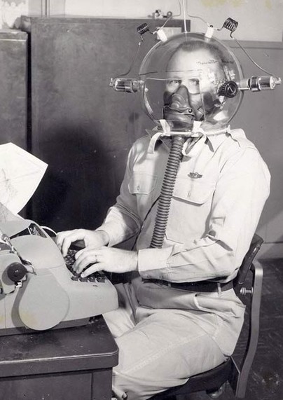 military person wearing some kind of strange helmet with electrodes on the outside of it, and a gas mask, sitting at a typewriter.