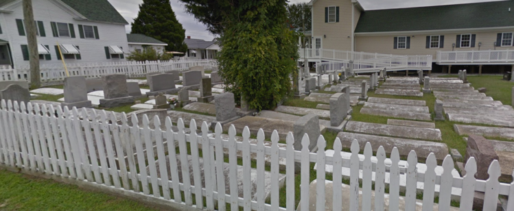via google street view, overlooking a cemetery lined with dozens of curved concrete forms in the shape and size of a casket, some with adjacent headstones, with a white picket fence along the edges and a plain wood construction church with nondescript 6