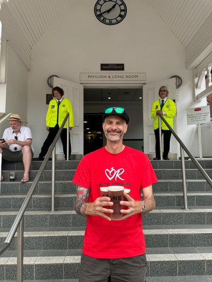 A man in a red t-shirt and cricket cap at the bottom of the steps leading into the members’ pavilion at Trent Bridge cricket ground. He’s holding 3 pints of King Mango IPA from the members’ bar. He’s flanked by two lovely stewards and is looking generally very happy with life. 