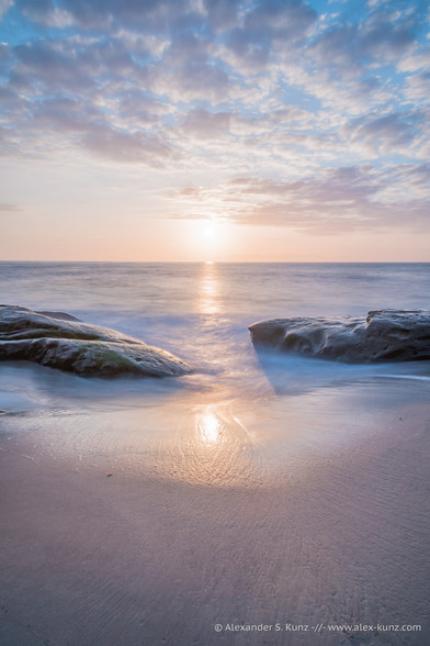 A vertical color seascape photo with smooth sand at the bottom, two coastal rocks left and right with a gap in between them through which the surf washes onto wet sand. The sun is sligthly above the horizon for sunset, and a few fluffy clouds ornament the sky.