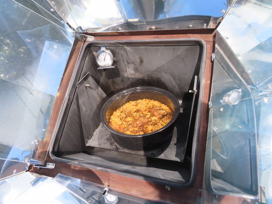 Global Sun Oven with reflectors. The glass lid is open and the lid on the pot is open, revealing birria, pulses, and grains. The thermometer shows a temp a bit below 300F.