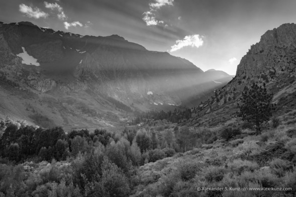 Black and white photo of a hazy mountain scenery in which a valley with trees at the valley floor is surrounded by steep rocky cliffs. Low afternoon light funnels through a pass and forms beams and rays where mountains and clouds block the light. 