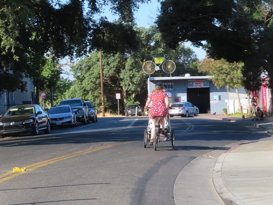 Woman wearing a short-sleeved lightweight print fabric summer dress while riding a trike that has a plastic bin in front in-between the two wheels. Ahead of her is a giant bicycle built to be a sign for a bike rental business. The entrance to the cycle path is just around the corner.