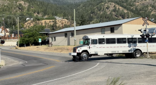 White bus with metal rail wheels inside of its rubber tires. The bus is slightly elevated up because there don't seem to be matching rail wheels on the back. It's crossing a street at a railroad crossing in Lillooet, British Columbia.