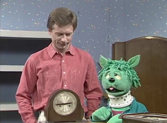 Mooncat, a green cat puppet with crosseyes and unruly hair (?) sits at a table next to a man with red hair in a pink shirt, and a wooden cased table clock.
