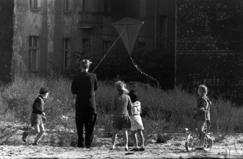 Photo of a (young man?) male figure flying an old fashioned kite at low altitude over a vacant lot in a city, with children watching.