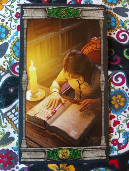 Card from the Vampires Tarot of the Eternal Night deck. 
Eight of Pentacles.
A man is seated at a table, leaning in studying a thick book volume. There is a bookmark on top of one page, and he is very focused. A lit candle to his right to provide light. Book shelves with books nearby to his right.