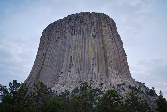 A massive, sheer rock formation known as a monolith, rising dramatically from a forested area. The rock surface features vertical striations and columns, giving it a distinctive, rugged appearance. The sky above is overcast with a soft, muted light.