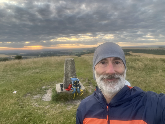 Selfie of me in front of the trig point on the top of Beacon Hill in southern England. At the foot of the trig point is my blue running pack and my Jetboil stove. 
